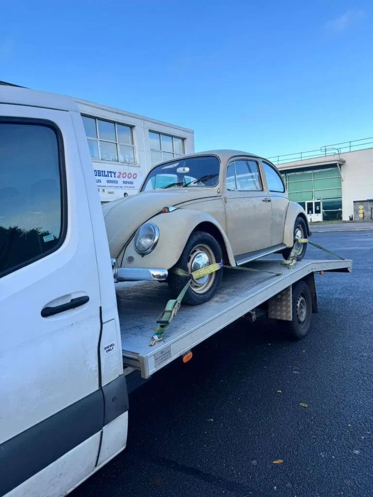 A white recovery truck transporting a classic vintage Volkswagen Beetle in an industrial area under a clear sky