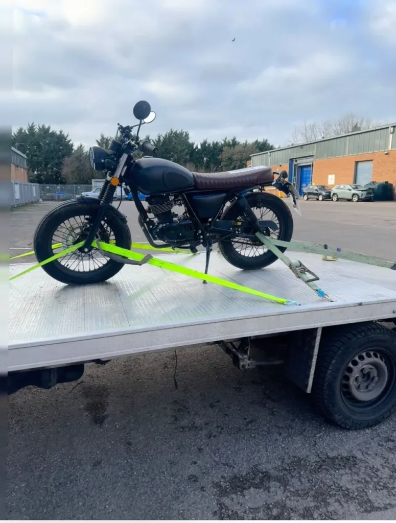 Black motorcycle secured on a recovery truck near a warehouse building.