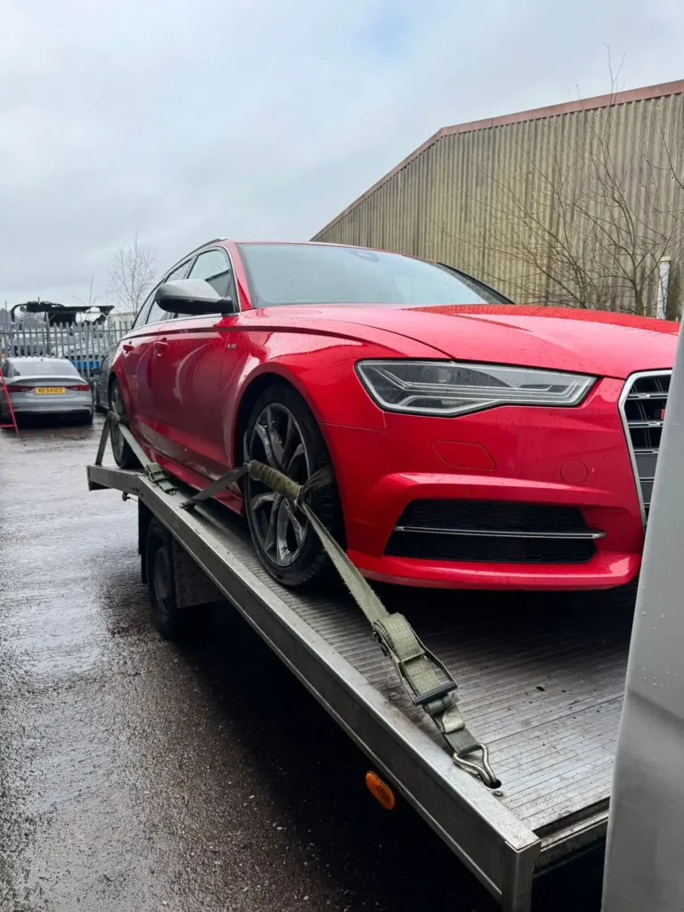 Red Audi sedan securely fastened on a recovery truck in a commercial yard area.