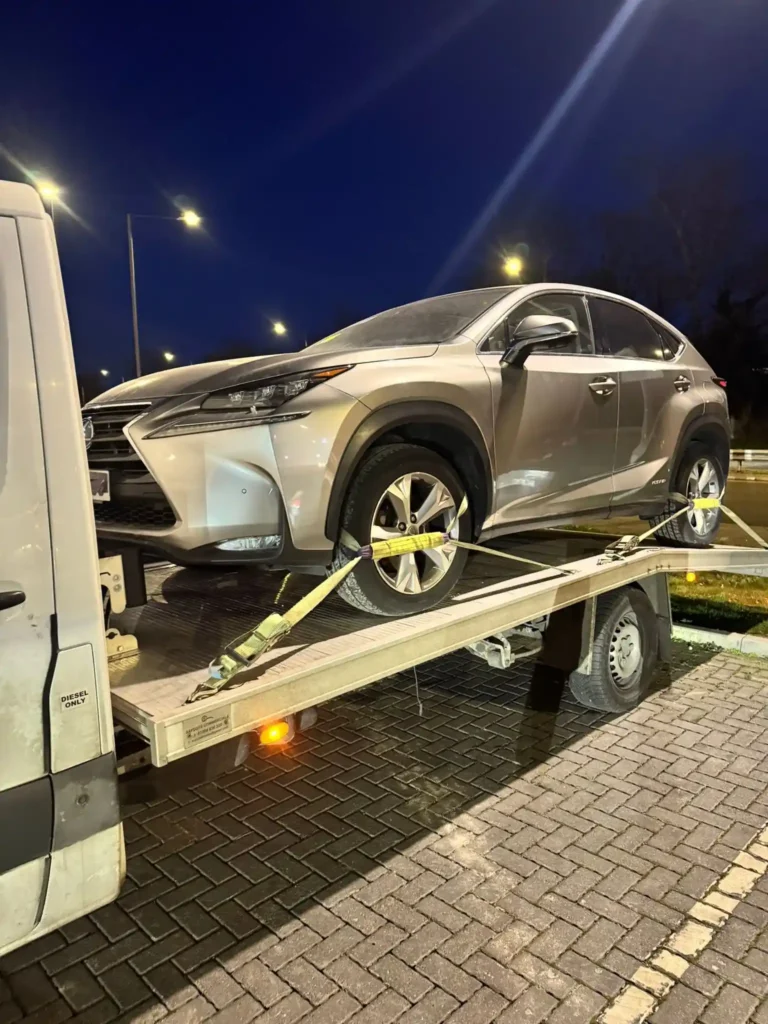 Silver Lexus SUV strapped on a recovery truck during night time in a lit parking area.