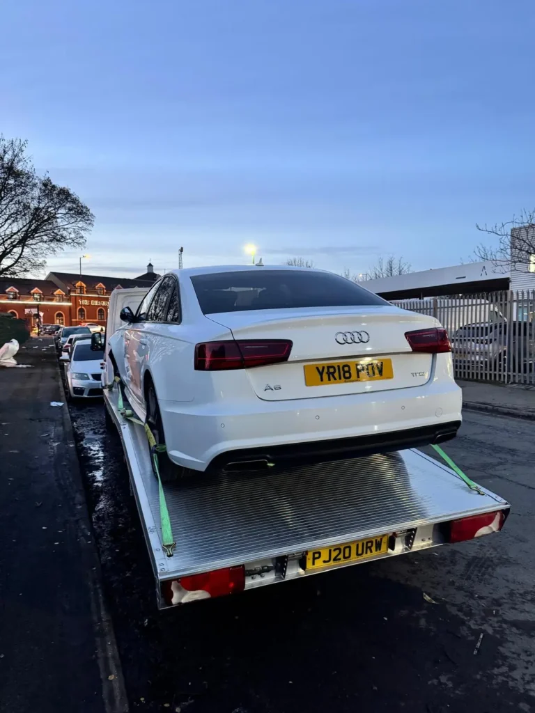 White Audi A6 secured on a recovery truck in front of a red-brick building